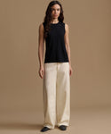 A woman stands against a plain warm beige studio background wearing a sleeveless black top and wide-leg cream pants.