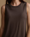 A close-up of a model wearing a sleeveless crew-neck tank top in a dark brown color, set against a solid warm beige background.