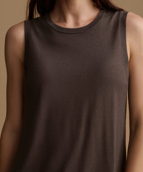 A close-up of a model wearing a sleeveless crew-neck tank top in a dark brown color, set against a solid warm beige background.