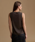 A woman shown from the back wearing a sleeveless dark brown knit top and a dark pleated skirt, set against a solid warm beige studio background.