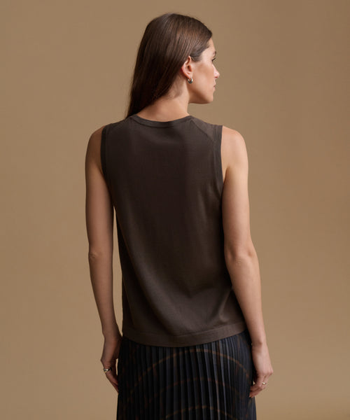 A woman shown from the back wearing a sleeveless dark brown knit top and a dark pleated skirt, set against a solid warm beige studio background.