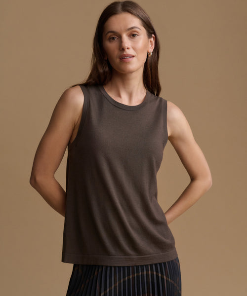 A woman modeling a sleeveless crew-neck tank top in a dark taupe-brown color, paired with a dark pleated skirt, against a solid warm beige studio background.
