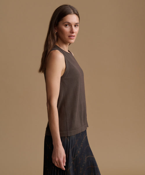 A woman wearing a sleeveless dark brown top and a dark patterned skirt, standing against a solid beige studio background.