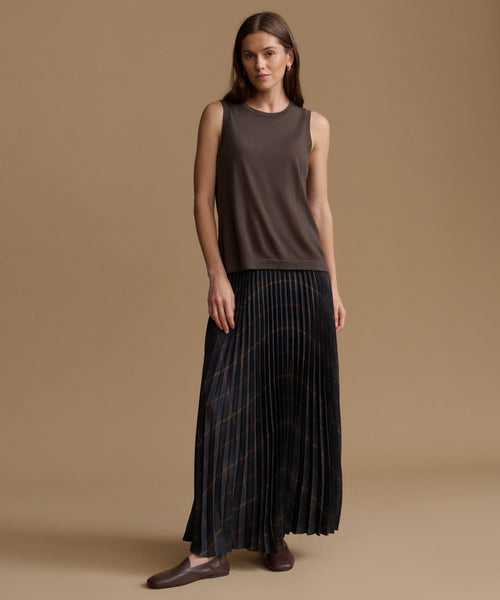 A woman modeling a sleeveless dark brown top and a long pleated skirt with thin gold stripes, standing against a solid tan studio background.