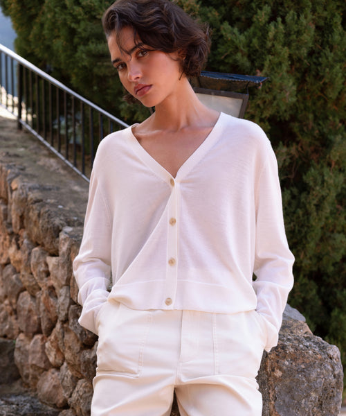 A person with short, wavy brown hair stands outdoors, leaning against a stone wall. They wear the Mira Cardigan with matching pants, set against greenery and a metal railing in the background.