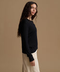 A woman modeling a black long-sleeve knit top with delicate lace-up detailing at the neckline, paired with cream-colored pants, set against a solid warm beige studio background.