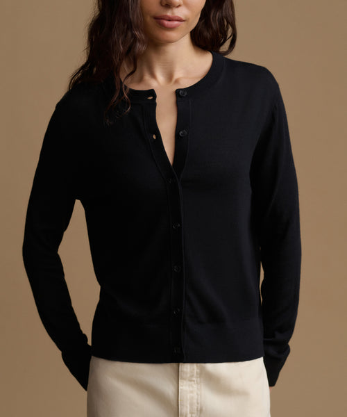 A woman wearing a black long-sleeve button-front cardigan with a relaxed fit, paired with light beige pants, set against a solid warm brown background.
