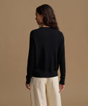 A woman shown from the back wearing a black long-sleeve knit top and cream wide-leg trousers, standing against a solid warm beige studio background.