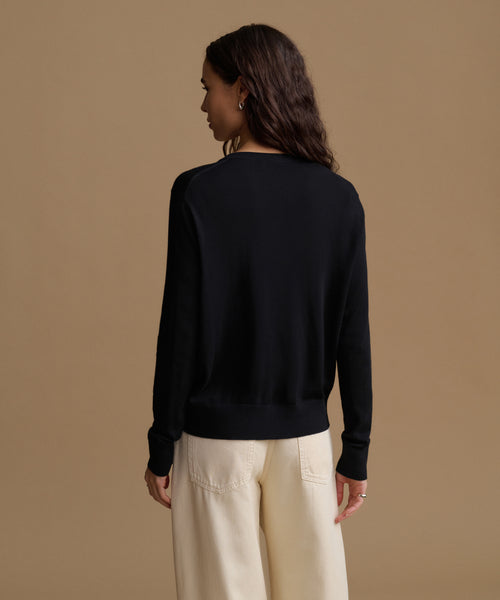 A woman shown from the back wearing a black long-sleeve knit top and cream wide-leg trousers, standing against a solid warm beige studio background.