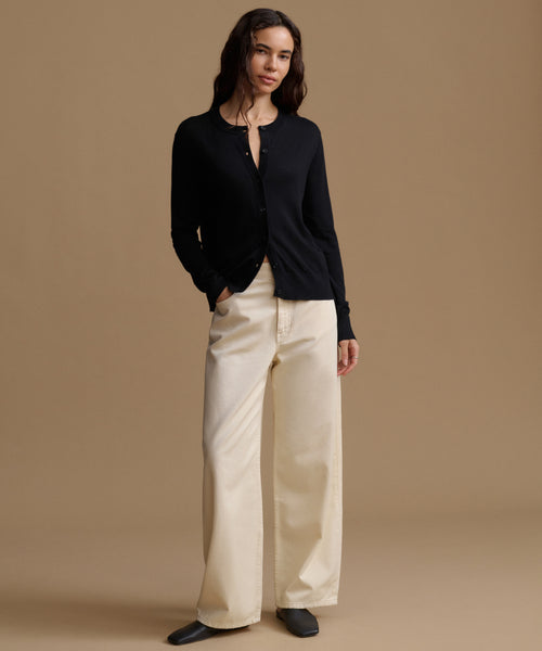 A woman modeling a black long-sleeve button-front cardigan with cream wide-leg trousers, standing against a solid warm beige studio background.