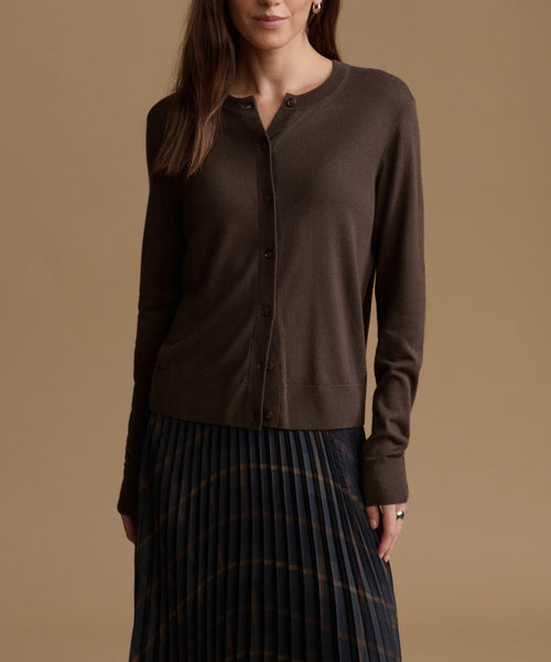 A woman wearing a dark brown long-sleeve button-front cardigan and a dark pleated skirt, standing against a solid warm tan studio background.