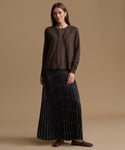 A woman stands against a warm beige studio background wearing a dark brown cardigan over a matching top and a long pleated navy skirt with subtle gold-toned stripes, paired with brown loafers.