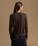 A woman shown from the back wearing a dark brown long-sleeve knit sweater and a dark pleated skirt, set against a solid warm beige studio background.