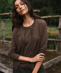 A woman wearing a dark brown button-front long-sleeve henley cardigan stands outdoors on a weathered wooden bridge with a green forest and water in the background.