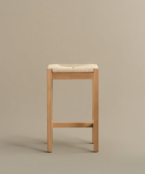 The Montecito Counter Stool features a minimalist design with straight wooden legs and a woven Danish paper cord seat, shown here in a Warm Oak finish against a plain beige background.