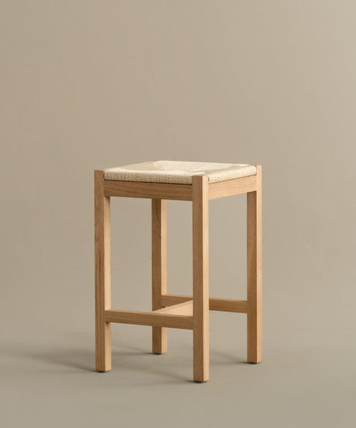 A Montecito Counter Stool featuring four warm oak legs and a woven Danish paper cord seat stands against a plain beige background.