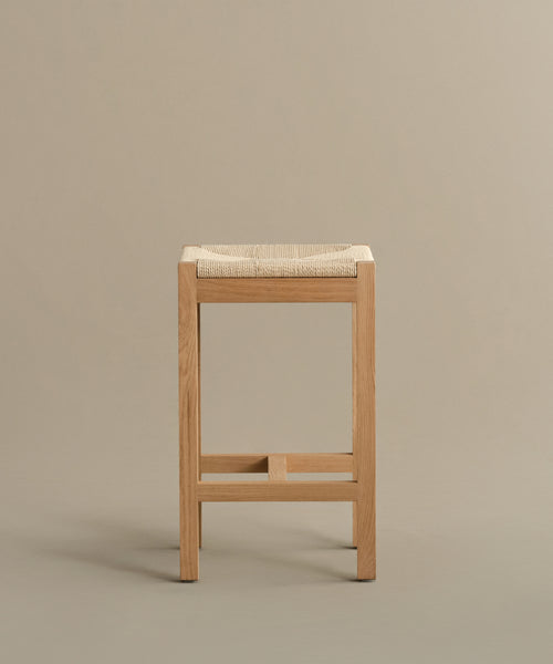 The Montecito Counter Stool features clean lines and a square Danish paper cord seat in warm oak, shown against a plain beige background. Its natural wood finish creates a minimalist, modern look.