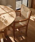 A Montecito Dining Arm Chair with a Danish paper cord seat and backrest is placed beside a round wooden table on a sunlit Warm Oak floor, with soft window and plant shadows creating an inviting atmosphere.
