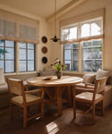 A cozy dining nook with a round warm oak table, three Montecito Dining Arm Chairs, built-in bench seating with cushions, a vase of flowers, and large windows with light filtered through linen shades.