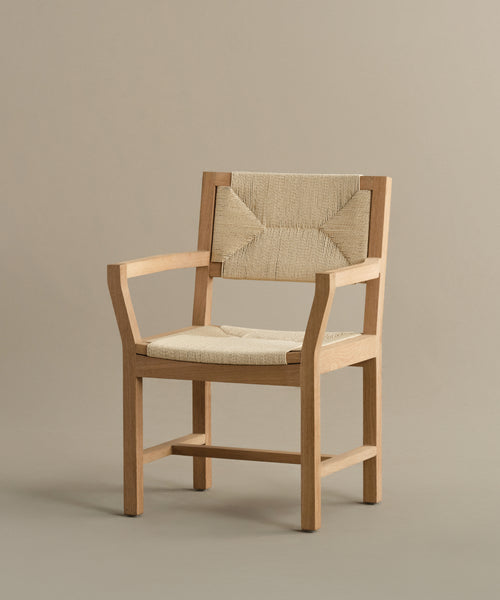 The Montecito Dining Arm Chair showcases a natural warm oak finish with beige Danish paper cord seat and backrest panels, all highlighted against a plain taupe background.