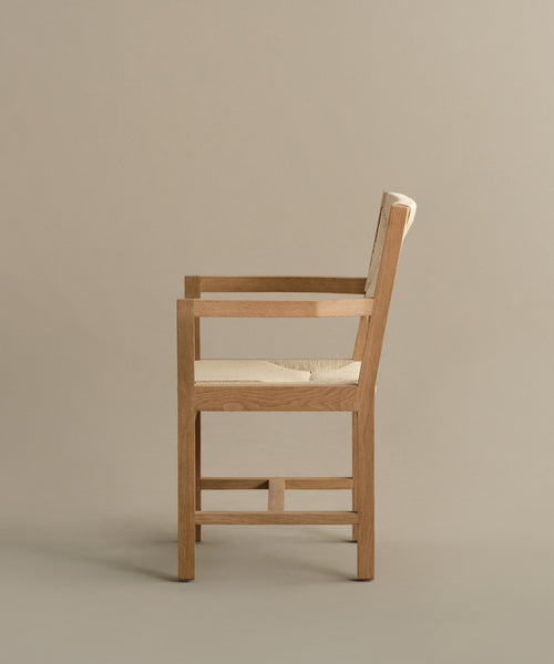 The Montecito Dining Arm Chair is displayed in profile against a neutral beige background, featuring a simple modern design with straight lines and a woven Danish paper cord seat and backrest. The finish shown is Warm Oak.