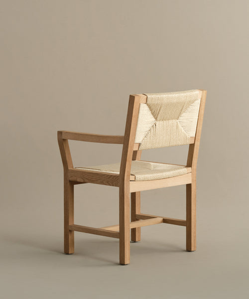 A minimalist Montecito Dining Arm Chair with clean lines, a warm oak natural finish, and woven Danish paper cord seat and backrest is angled slightly to the left against a plain beige background.