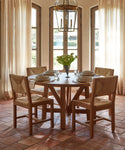 A round wooden dining table for four is surrounded by Montecito Dining Chairs in Warm Oak with handwoven Danish paper cord seats, set on a tiled floor with light streaming through tall windows and a vase of flowers on the table.