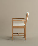 Side view of the Montecito Upholstered Dining Arm Chair with a light-colored cushioned seat and backrest, clean straight lines, and minimalist white oak design, set against a plain beige background.