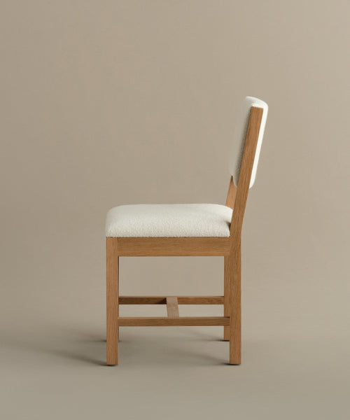 A side view of the Montecito Upholstered Dining Chair, featuring a cushioned seat and backrest in soft ivory boucle, set against a neutral beige background.