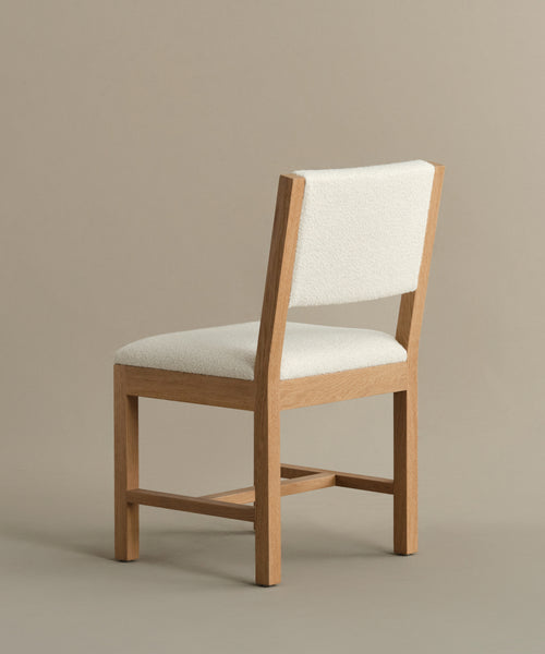 The Montecito Upholstered Dining Chair features a simple white oak frame with ivory boucle seat and backrest, shown on a neutral beige background at an angle to highlight both its side and back.