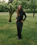 A woman stands in a grassy park with trees wearing a black long-sleeve and black skirt and black ankle boots.