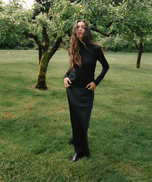 A woman stands in a grassy park with trees wearing a black long-sleeve and black skirt and black ankle boots.