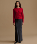 A woman stands against a plain warm beige studio background wearing a bright red long-sleeve button-front cardigan and a long dark charcoal skirt.