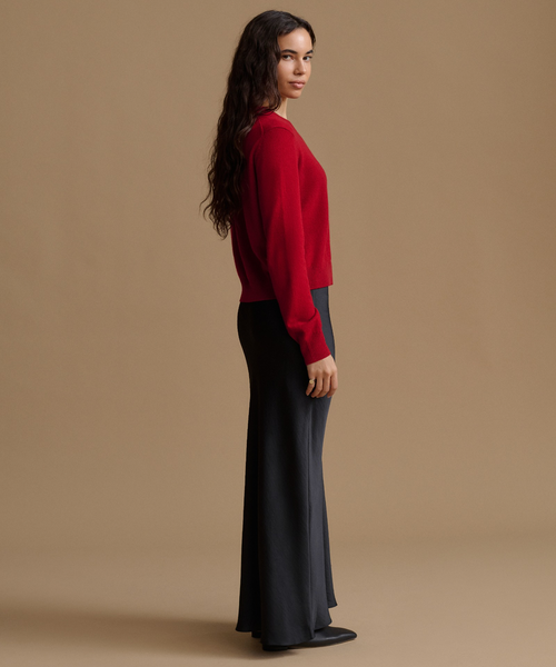 A woman stands in profile wearing a bright red long-sleeve knit top and dark charcoal wide-leg trousers against a plain warm beige studio background.