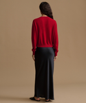 A woman is shown from behind wearing a bright red knit sweater and a long black satin skirt against a solid tan studio background.