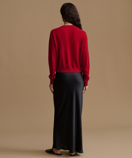 A woman is shown from behind wearing a bright red knit sweater and a long black satin skirt against a solid tan studio background.