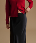 A model wearing a vibrant red button-up cardigan and a black satin midi skirt, shown from the chest down against a solid beige background.