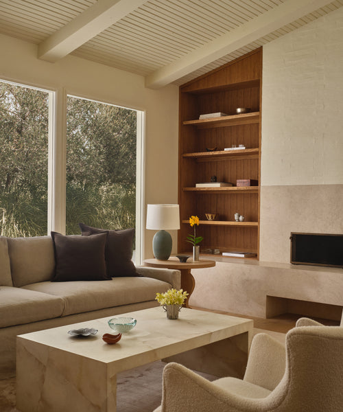 A cozy, neutral-toned living room features large windows, a beige sofa with dark pillows, two cream chairs, built-in wooden shelves, a fireplace, and an ivory Napa Coffee Table with decor. Sunlight fills the inviting space.