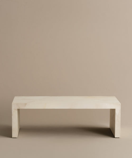 The Napa Coffee Table features a minimalist, rectangular design in a light, smooth finish with clean lines, reminiscent of ivory. It is set against a plain, neutral beige background.