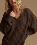 A woman wearing an oversized dark brown cable-knit V-neck sweater against a warm beige background.
