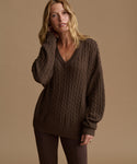 A woman modeling an oversized brown cable-knit V-neck sweater against a solid warm beige studio background.