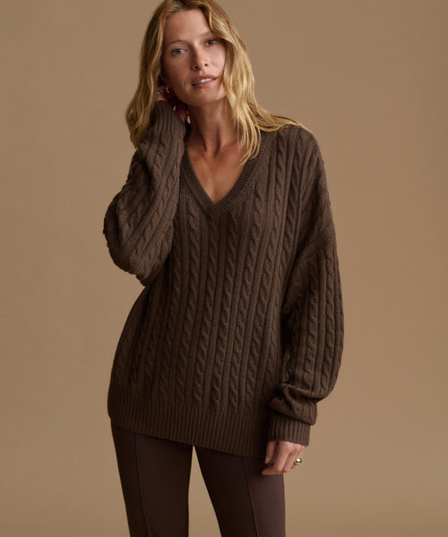 A woman modeling an oversized brown cable-knit V-neck sweater against a solid warm beige studio background.