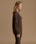 A woman in side profile wearing a dark brown cable-knit sweater and matching dark brown pants, standing against a solid warm beige studio background.