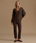 A woman stands against a plain warm beige studio background wearing a dark brown cable-knit V-neck sweater, matching dark brown trousers, and dark brown loafers.
