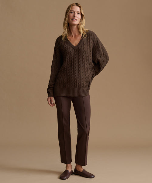 A woman stands against a plain warm beige studio background wearing a dark brown cable-knit V-neck sweater, matching dark brown trousers, and dark brown loafers.