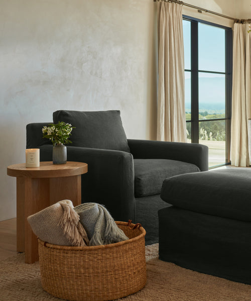 A cozy living room corner features a dark armchair, ottoman, wooden side table with a plant and candle, and a Nesting Basket filled with folded blankets near large windows dressed in beige curtains.