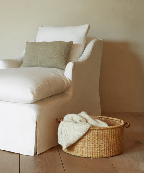 A white upholstered armchair with a beige cushion sits on wooden flooring. Beside it, a Nesting Basket holds a folded cream knit blanket. The scene features a plain, light-colored wall in the background.