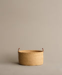 The Nesting Basket, a round handwoven elephant grass basket with brown handles, rests on a plain light beige surface against a matching beige background.