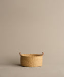 A Nesting Basket made from handwoven elephant grass, featuring two small brown handles, is centered and empty on a plain light beige background. Crafted by Ghanaian artisans.
