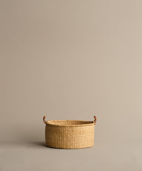 A Nesting Basket made from handwoven elephant grass, featuring two small brown handles, is centered and empty on a plain light beige background. Crafted by Ghanaian artisans.